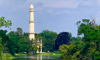 Ein alleine dastehendes hohes Minarett inmitten eines Parks vor einem Teich.