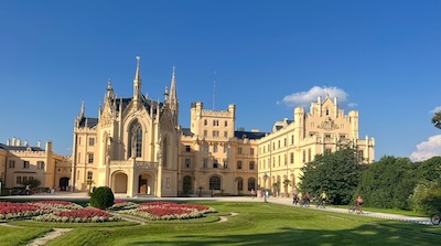Fassade eines Schlosses der Neo-Tudorgotik, davor eine grüne Wiese mit Blumenbeet.