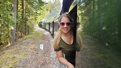 Blonde Frau mit Sonnenbrille schaut bei der Waldviertelbahn aus dem Zug heraus - im Hintergrund der Wald, die Strecke und die Dampflok mit den grünen Waggons