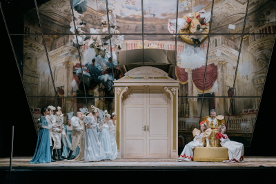 Spiegelfläche über dem Bühnenboden eines Theaters, Spiegelung zeigt Fassade des Schönbrunner-Theatersaals, auf der Bühne selbst: Darsteller in hellen Gewändern (blau, weiß), auf der rechten Bühnenseite: eine Sitzgarnitur in Epaulettenform