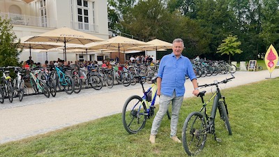 Ein Mann mit hellblauem Hemd stützt sich auf ein Fahrrad. Dahinter ein großer voll besetzte Radständer, dahinter Sonnenschirme eines Gastgartens.