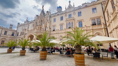 Palmen im Pflanzkübel und Sonnenschirme in einem Gastgarten vor einer neugotischen Schlossfassade.