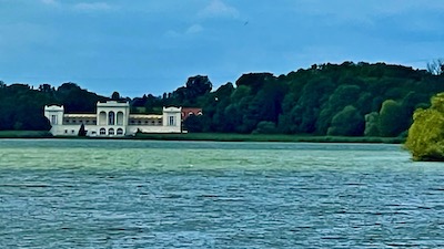Ein klassizistisches kleines Schloss vor einem großen Teich. Im Hintergrund Bäume.