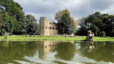 Eine Burgruine in einem Park. Davor eine Wiese und eine Wasserfläche, in der sich die Burgruine spiegelt.