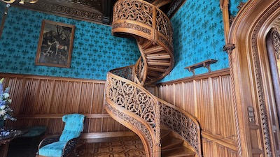 Eine holzgeschnitzte Wendeltreppe vor einer holzvertäfelten und blau tapezierten Wand.