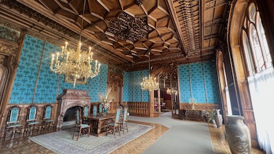 Ein mit einer türkisten Stofftapete ausgestatteter neugotischer Schlossraum mit einem Messingluster und einer Holzkassettendecke.