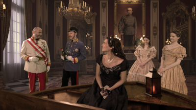 Screenshot aus einer virtuellen 5D-Reise zeigt Sisi-Darstellerin in einem schwarzen Kleid in einem Bootsitzend, das im Prunkraum eines barocken Palasts Halt macht...links von SIsi: Kaiser Franz Josepf in rot-weißer Uniform, neben im Kronprinz Rudolf in grau-schwarz-roter Uniform, rechts von Sisi: ihre Töchter Gisela und Marie Valerie in beige-rose-farbenen Kleidern...hinter ihnen: ein Gemälde von Kaiser Franz Joseph