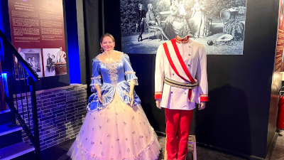 zwei kopflose Statuen...einmal in einem blau-weißen kleid, einmal in einer rot-weißen Uniform...vor der Statue im Kleid steht eine Frau, die ihren Kopf am Halsansatz der Statue anlegt...dahinter Informationstafeln und Bilder von den Habsburgern
