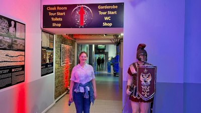 Frau in weißer Bluse und blauer Jeans steht vor dem Eingang im Time Travel Vienna, zu ihrer Rechten: die Statue eines römischen Soldaten mit Schild und Helm....zu ihrer Linken: Informationstafeln über das antike Wien...hinter ihr: ein Schild mit der Aufschrift "Garderobe, Tour Start, WC, Shop"