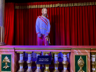 eine Wachsfigur von Kaiser Franz Joseph in weiß roter Uniform vor einem rot-goldenen Vorhang...davor ein Balkon in Grün, Silber und Marmor mit einer Tafel, die Geburts-und Todesdaten des Habsburgers angibt