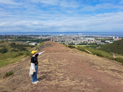 Eine Frau steht am Arthurs Seat und zeigt auf Edinburgh.