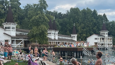 Weiß gestrichen Badehäuser aus Holz am Hévízer Thermalsee. Davor Stege und zahlreiche Badegäste.