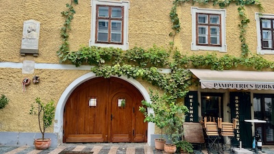 Fassade eines alten, crèmegelb gestrichenen Hauses, mit bogenförmigem Tor, einem Gassenlokal und einer Gedenktafel.