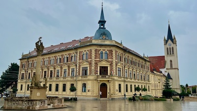 Ein großes gründerzeitliches Gebäude mit einem Mittelturm, dahinter der neugotsiche Turm einer Kirche.