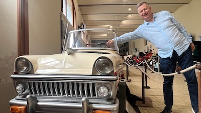 Ein kleines Auto, mit dem auch Kinder auf einem geschützten Gelände fahren können. Daneben ein Mann in hellblauem Hemd.