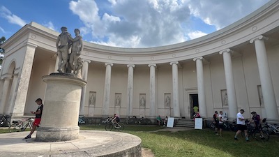 Ein pseudo-griechischer Rundtempel mit zahlreichen Säulen vor einem Arkadengang, in weiß gestrichen. Davor auf einem Podest eine Gruppe von drei weiblichen Figuren. Viele Radfahrer, darunter einer, der an der Podestsäule lehnt