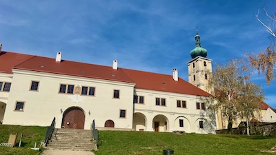 Herrenhaus Draškovič in Čachtice und die Pfarrkirche von Čachtice. Renaissancegebäude mit weißer Fassade vor grüner Wiese, dahinter ein Kirchturm.