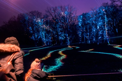 Mutter und Kind vor den Singing Lights - bunte Lichterstrahlen schlängeln sich bis zum Wald, der violett beleuchtet wird.