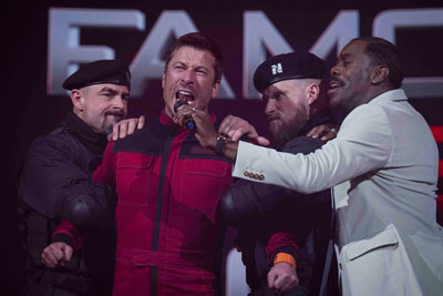 Glen Powell wird von Soldaten gehalten, während er in ein Mikrofon brüllt, das der böse Gameshow-Host hält