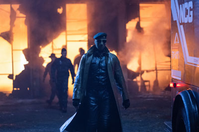 Ein Soldat mit Maske und Sonnenbrille bewegt sich in The Running Man von einem brennenden Gebäude weg