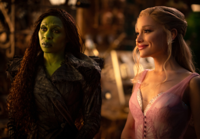 Elphaba und Glinda. Elphaba hat grüne Haut und ein schwarzes Kleid. Glinda hat blonde Haare und ein pinkes Kleid.