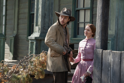 Mann in beiger Jacke und Hut, Frau in rosa Kleid vor Haus