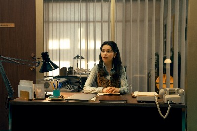 Frau mit langen dunklen Haaren, weiße Bluse mit braunem, ärmellosen Sweater, in einem Büro sitzend