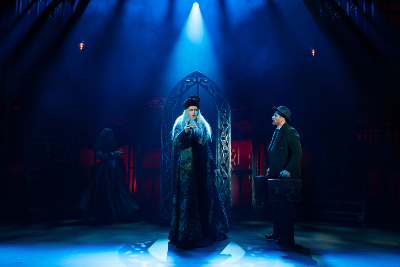 Drei Figuren auf einer Bühne, dunkelblaues Licht, abgespitzter Arkadenbogen im Huntergrund..in der Mitte: Mann in dunkler Kleidung mit weißem Pelz, rechts Mann mit Aktenkoffer und dunklem Anzug, links: Frau in Kleid