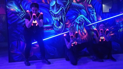 Drei Lasertag Spielerinnen posieren vor einer Wand mit Alien in blau-rotem Neonlicht.