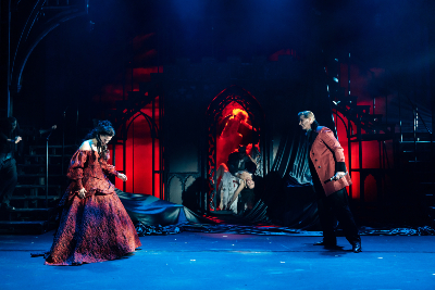 Bühnenbild in rötlich-blauem Licht...Mann und Frau in rötlicher Kleidung gegenüberstehend..in der Mitte: Frauenkörper in einem rot erleuchteten Durchgang