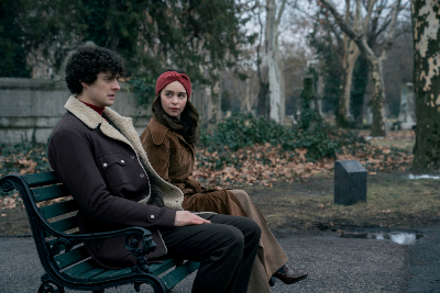 Mann mit schwarzen lockigen Haaren und schwarzer Jacke und weißem Revers neben Frau mit rotem Stirnband und braunem Mantel auf Parkbank sitzend