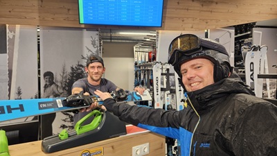 Lächelnder Skifahrer holt sich Leihski, die ihm der Mitarbeiter beim Skiverleih Mönichkirchen über den Tresen reicht.