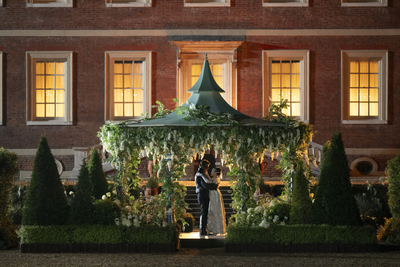 eine Art Gazebo in Grün, umringt von Pflanzen und Gartenhecken bei Nacht, darunter ein maskiertes, tanzendes Pärchen.....Gazebo befindet sich vor Ziegelsteinhaus mit hell erleuchteten Fenstern