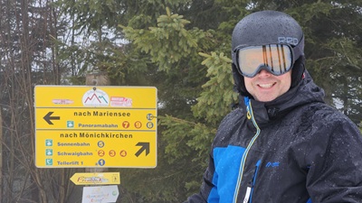 Lächelnder Mann mit Skihelm und Brille vor dem Schild bei dem die Pisten nach Mariensee oder Mönichkirchen abzweigen.