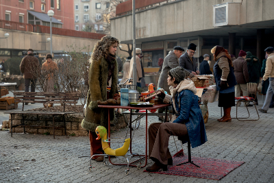 Menschen auf einem Flohmarkt, Frau in einem braunen Mantel vor einem TIsch stehend, dahinter andere Frau in blauer Jacke, grauer Haube und brauner Hose, neben ihr eine Plastikente, Bänke und rote sowie weiße Gebäude im Ostblock-Stil
