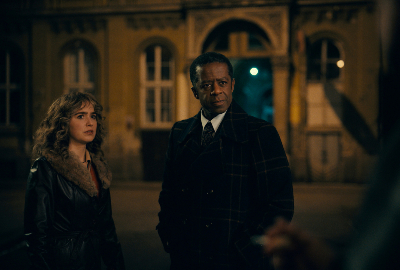 Frau mit dunkelblonden gewellten Haaren in brauner Jacke neben afroamerikanischem Mann in einem schwarzen ANzug, hinter ihnen: eine Gebäudefassade bei Nacht