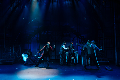 Bühnenbild in blauem Licht...Gruppe von Männern, einer hält ein Kreuz in der Hand...gegenüber von ihm: Mann wie ein Vampir gekleidet in rotem Anzug mit blutverschmiertem Mund