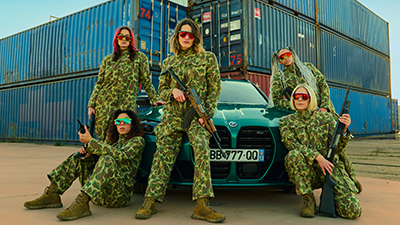 Fünf Frauen in Militäranzügen mit Waffen posen vor einem Auto und Schiffscontainern