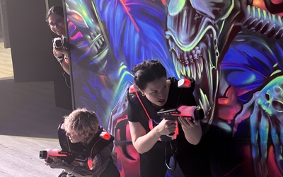 3 Frauen spielen bei LaserDance im Prater, eine lächelt hinter einer Wand, die ein Alien zeigt.