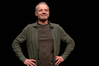 Thomas Maurer grinsend, in khaki-farbener Jacke und braunem Shirt, die Hände gegen die Hüften gestemmt