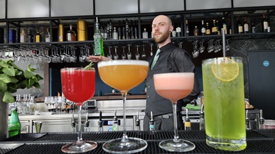 Die Cocktails The Flaming Botanist, Pornstar Martini, Pink Devil und Tokyo Iced Tea in der Q27 Bar in Linz, dahinter balanciert der Barchef eine Flasche auf der Hand.