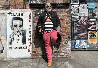 Christian Orou in der Arena - Mann mit langen grauen Haaren, Sonnenbrille und rosa Hose lehnt an einer Ziegelwand neben einem The Clash Poster