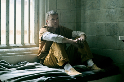 Mann in orangenem Gefängnisjumpsuit auf einer Pritsche sitzend... gespielt von Sam Worthington.