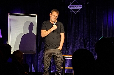 Andreas Ferner mit Mikro in der Hand vor Flipchart / Tafel stehend, eine Hand in der Hosentasche