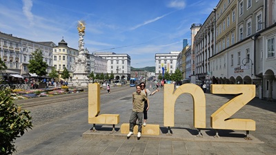 Pärchen steht beim I des Linz Schriftzugs am Hauptplatz von Linz. Im Hintergrund die Dreifaltigkeitssäule