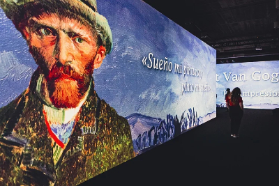 Großformatige immersive Ausstellung mit einer Projektion von Vincent van Goghs Selbstporträt auf einer langen Wand. Daneben ist ein spanisches Zitat eingeblendet, während eine einzelne Besucherin im abgedunkelten Raum die Installation betrachtet.
