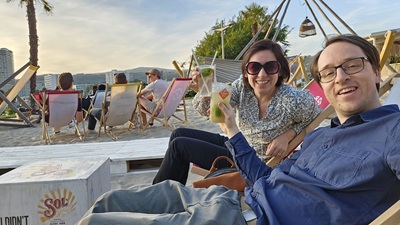 Mann und Frau sitzen in Liegestühlen in der Sandburg Bar und stoßen mit Cocktails an.
