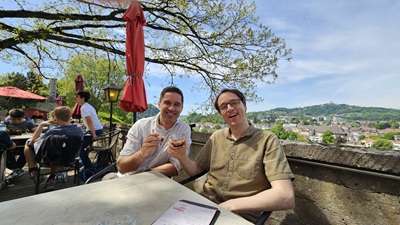 Zwei Männer stoßen im Schloss Café in Linz an - im Hintergrund der Blick auf die Stadt.
