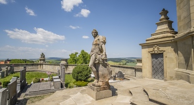 Barockfriedhof in Střílky: Eine barocke Engelsstatue im Vordergrund, dahinter eine barocke Friedhofsmauer mit Skulpturen.
