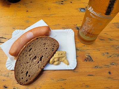 Knackwurst, Brotscheibe und Senf auf einem Pappteller, daneben eine Flasche Almdudler auf einem Heurigentisch